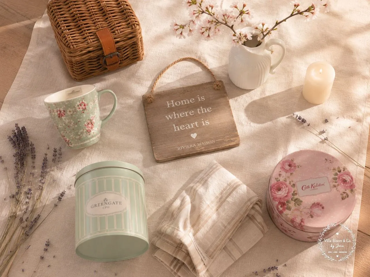 Flatlay mit GreenGate Floral-Tasse, GreenGate Vorratsdose mint, Riviera Maison Holzschild Home is where the heart is, Cath Kidston Rosen-Dose, Korb mit Lederlasche, Kirschblütenzweige, Leinen und Lavendel